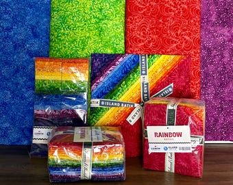 May include: A collection of colorful fabric bundles, including rainbow-themed packs and patterned fabrics in red, green, blue, and purple. The bundles are wrapped and labeled with the "Island Batik" brand.