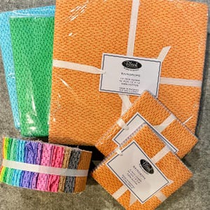 May include: Stacks of fabric squares in various colors, including orange, blue, green, and a rainbow assortment. The orange fabric is labeled "Raindrops" and is 100% cotton. The squares are tied with white ribbon, perfect for quilting or crafting projects.