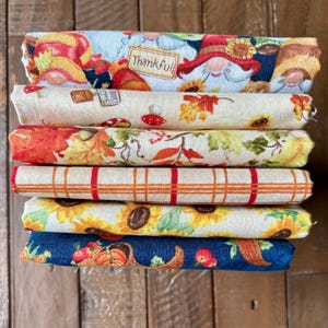 May include: A stack of six rolled fabric pieces with autumn-themed patterns. Designs include gnomes, sunflowers, fall leaves, and plaid. One piece has the word "Thankful" on it. The fabrics are in shades of blue, orange, yellow, and red.