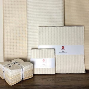 May include: A collection of Latte Andover Fabrics in various sizes and patterns. The fabrics feature cream and beige tones with striped and dotted designs. Some are bundled and tied with a ribbon, while others are stacked. The Andover Fabrics label is visible.