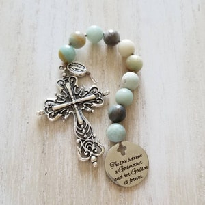 May include: A silver cross charm with a beaded rosary chain made of light blue, grey, and white stones. The charm has a small silver pendant with the Virgin Mary on it. The rosary chain has a silver tag with the text "The love between a Godmother and her Godson is forever".