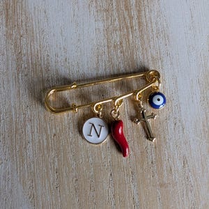 May include: Gold-coloured safety pin charm with dangling pendants. Charms include a white circle with the letter "N", a red chilli pepper, a gold cross, and a blue evil eye. The pin is displayed on a light wood surface.