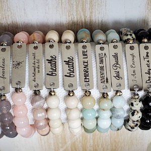 Könnte beinhalten: Zehn Armbänder mit verschiedenen bunten Perlen und Metalletiketten mit inspirierenden Sprüchen. Die Sprüche lauten: inhale exhale, ...fearless, she believed she could... so she did, breathe, breathe, EMBRACE LIFE, ADVENTURE AWAITS, Just Breathe, I Am Enough, One day at a Time, Shine Bright.