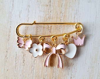 Può includere: Una spilla a forma di spillo da balia dorata con ciondoli rosa e bianchi. I ciondoli includono una farfalla, un fiore, un fiocco, un cuore e una corona. La spilla è un accessorio decorativo.