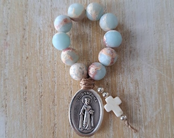 May include: A light blue and white stone rosary with a silver charm featuring a depiction of St. Dymphna. The rosary is tied with a brown cord and has a white cross charm.