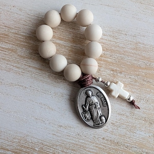 May include: A handcrafted rosary with large, pale wooden beads. It features a silver-toned St. Mark medal and a small white cross. The beads are strung together with a brown cord.