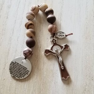 May include: A brown and white rosary with a silver crucifix and a silver medal featuring the Virgin Mary. The rosary beads are made of stone and have a marbled pattern. The crucifix has a brown wooden cross with a silver Jesus figure. The medal has a silver frame with a white image of the Virgin Mary. The rosary is hanging on a white wooden surface. The text on the medal reads "IN RI". The text on the back of the rosary reads "Our Father, who art in heaven, hallowed be thy name, thy kingdom come, thy will be done, on earth as it is in heaven. Give us this day our daily bread. And forgive us our debts, as we forgive our debtors. And lead us not into temptation, but deliver us from evil. For thine is the kingdom, the power and the glory for ever and ever. Amen."