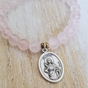 May include: A pink rose quartz bracelet with a silver charm featuring a depiction of Saint Lucy. The charm is inscribed with "St. Lucy" and "Italy".