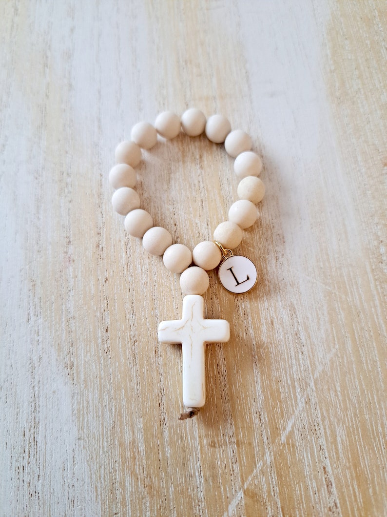 May include: A white wooden beaded bracelet with a white cross charm and a gold charm with the letter "L" on it.