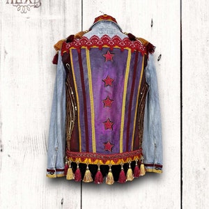 May include: A denim jacket with a purple, red, and gold striped back panel. The panel has four red stars and a gold trim. The jacket has red tassels on the bottom.