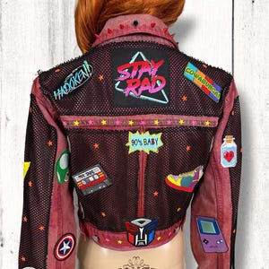 Puede incluir: Una chaqueta vaquera roja con un panel de malla y varios parches, incluyendo "Stay Rad", "Cowabunga", "90's Baby", "Hadoken", "Mix Tape", "Dope", "Empire Strikes Back" y una Game Boy.