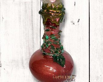 May include: A decorative bottle with a red, glittery finish. The bottle is adorned with green leaf-shaped embellishments and a clear, iridescent stopper. The neck of the bottle is accented with gold and green paint.