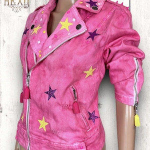 Può includere: Giacca di jeans rosa con decorazioni a forma di stella in giallo, viola e bianco. La giacca ha una chiusura a zip, due tasche e un colletto con borchie.
