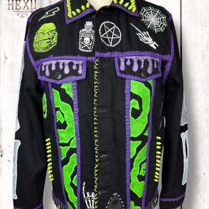 Könnte beinhalten: Schwarze Jeansjacke mit neongrünen und lila Akzenten, bestickt mit Aufnähern eines Wesens, eines Pentagramms und eines Spinnennetzes. Die Jacke hat eine Naht auf der Vorderseite und Skelettknochen-Designs auf den Ärmeln. Der Kragen hat neongrüne Stacheln.