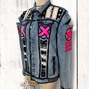 Pode incluir: Uma jaqueta jeans azul claro com lavagem ácida, acabamento preto, ombros recortados e painéis verticais entrelaçados com tiras brancas e pretas. A jaqueta é decorada com símbolos X rosa choque, um emblema de estrela e listras na manga.