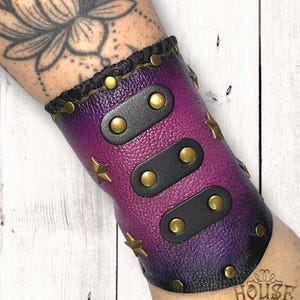 Puede incluir: Un brazalete de cuero morado y negro con detalles de estrellas y tachuelas doradas. El brazalete presenta correas de cuero negro y un borde trenzado. Un accesorio único.