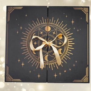 Puede incluir: Una caja negra con un diseño celestial dorado, con un sol, lunas y estrellas. La caja está atada con una cinta dorada. Las esquinas tienen detalles geométricos dorados. La caja está cerrada.