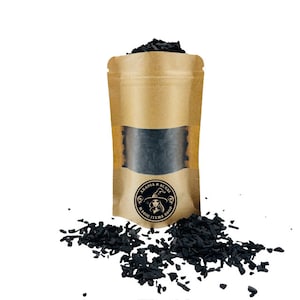 May include: A brown paper bag with a clear window, filled with black charcoal pieces. The bag features a logo that says "Abadia & Sense Magic Items Shop". Some charcoal pieces are scattered around the bag.