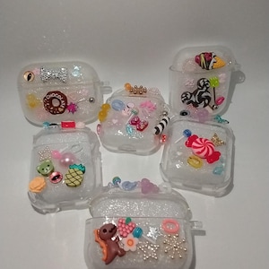May include: Clear AirPod cases decorated with various charms, including bows, donuts, candies, and bears. The cases are embellished with glitter and small beads, creating a playful and whimsical aesthetic. The cases are designed to protect and personalize Airpods.