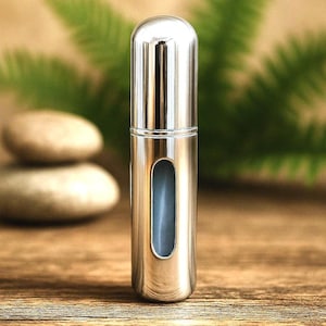 May include: A small, silver-colored perfume atomizer with a rounded top and a vertical, oval-shaped window. The atomizer is set against a blurred background of green foliage and a wooden surface.
