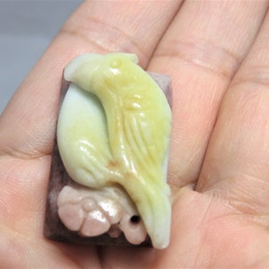 May include: A carved jade pendant featuring a bird with yellow and green tones, perched on a pink and purple base.