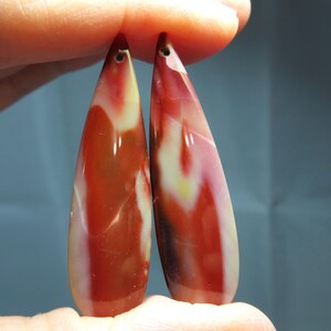 May include: Two teardrop-shaped pendants made of polished stone with a smooth, glossy finish. The stones feature a marbled pattern with vibrant swirls of deep red, cream, and hints of yellow, each with a small drilled hole at the top for hanging.