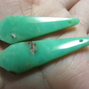 May include: Two polished, light green gemstone pendants. Each pendant has a pointed end and a hole drilled near the tip. The gemstones have a few small brown inclusions.