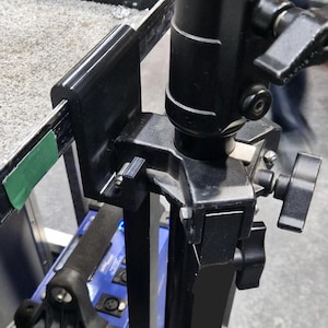 May include: A black metal stand with a clamp for attaching a light or camera. The stand is attached to a black and blue power source.