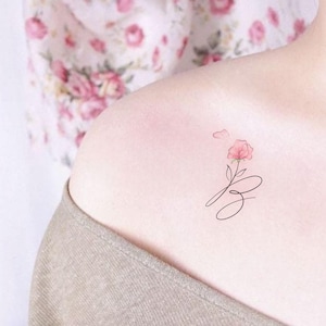 May include: A delicate tattoo design featuring a pink flower with a stem and leaves, and a cursive letter 'S' on the shoulder.