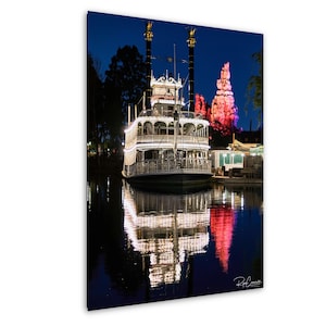 May include: A nighttime photograph of a steamboat on a body of water, with a mountain in the background. The steamboat is white with lights, and the water reflects the lights and the mountain. The sky is dark blue.