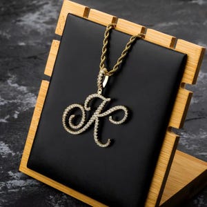 Peut inclure: Un collier en chaîne dorée avec un pendentif en forme de lettre K cursive serti de diamants. Le collier est présenté sur un présentoir en bois avec un revêtement en velours noir.