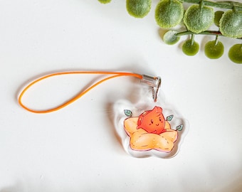 May include: A clear acrylic charm featuring a small, smiling red character nestled inside a light orange, star-shaped cushion. The charm is attached to a thin, bright orange elastic loop with a silver-toned metal connector.