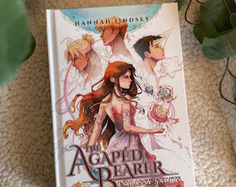May include: A hardcover book titled The Agaped Bearer Embassy Saints by Hannah Lindsey. The cover features an illustration of a central female figure with long brown hair surrounded by three other characters, all set against a white background.