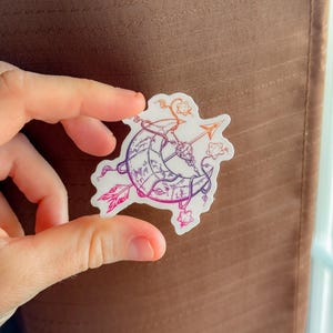 Op de afbeelding: Een sticker met een halve maan doorboord door een pijl, met bloemen en hemelse accenten. Het ontwerp maakt gebruik van een verloop van paarse, oranje en roze tinten. De sticker wordt tussen vingers gehouden tegen een bruine achtergrond.