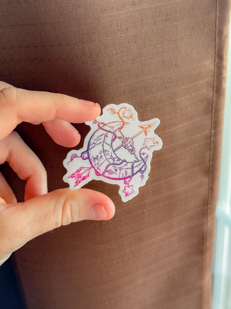 Op de afbeelding: Een sticker met een halve maan doorboord door een pijl, met bloemen en hemelse accenten. Het ontwerp maakt gebruik van een verloop van paarse, oranje en roze tinten. De sticker wordt tussen vingers gehouden tegen een bruine achtergrond.