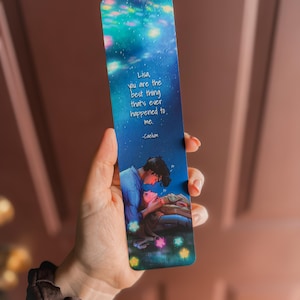 May include: A bookmark with a starry night sky design and a romantic illustration of two people. The text reads, "Lisa, you are the best thing that's ever happened to me." The bookmark is held in a hand.