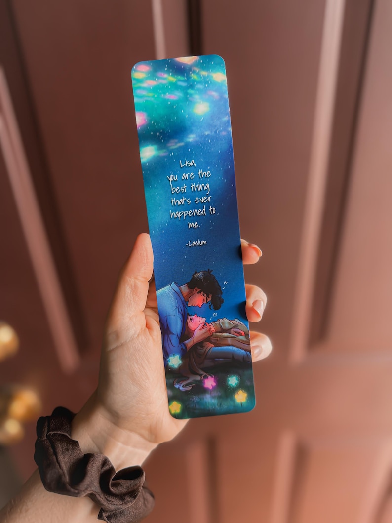 May include: A bookmark with a starry night sky design and a romantic illustration of two people. The text reads, "Lisa, you are the best thing that's ever happened to me." The bookmark is held in a hand.