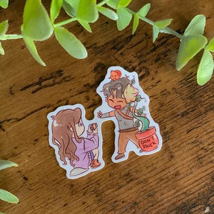Pode incluir: Um autocolante com duas personagens de desenhos animados. Uma figura está ajoelhada e a tirar uma foto, enquanto a outra segura uma planta num vaso com o texto "DON'T PLUCK". O autocolante tem uma borda branca e está sobre um fundo de madeira.