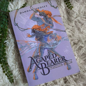 May include: A hardcover book titled "The Agaped Bearer Wishing Stars" by Hannah Lindsey. The cover features a dynamic illustration of two figures in shades of blue and orange against a lavender background. The book is resting on a white, textured surface.