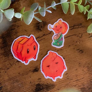 Könnte beinhalten: Drei orangefarbene Cartoon-Aufkleber auf einer Holzoberfläche. Einer ist ein würfelförmiger Charakter, ein anderer isst Chips, und der dritte hat einen traurigen Ausdruck. Grünes Laub ist im Hintergrund.