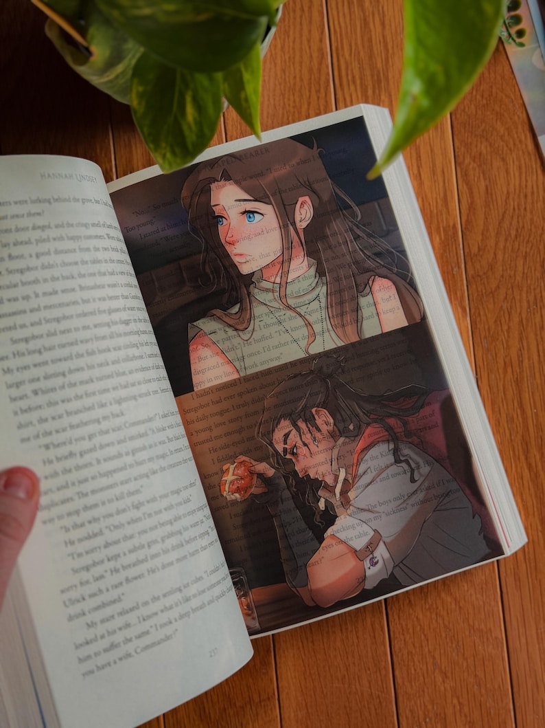 May include: Open book with illustrations. One illustration shows a woman with long brown hair and blue eyes. The other illustration shows a man with long dark hair. The book is open on a wooden surface.