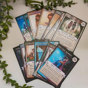 May include: A collection of fantasy-themed trading cards, featuring illustrations of characters and locations. The cards have a black border and include text describing each card's attributes. The cards are fanned out on a white surface.