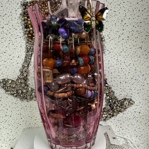 May include: A pink glass vase filled with various colorful beads, bracelets, and jewelry. A gold-toned butterfly-shaped clip is attached to the rim. The vase is on a white rotating stand, with additional jewelry visible in the background.