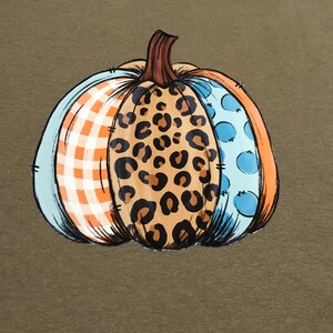 May include: A green t-shirt with a colorful pumpkin design. The pumpkin is made up of three different patterns: plaid, leopard print, and polka dots.
