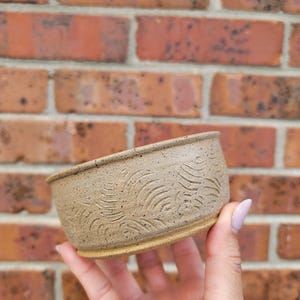 May include: A small, round, speckled beige ceramic bowl featuring a textured, carved pattern of repeating wave-like lines around the exterior. The bowl has a smooth, slightly flared rim and a flat base, showcasing a natural, earthy clay finish.