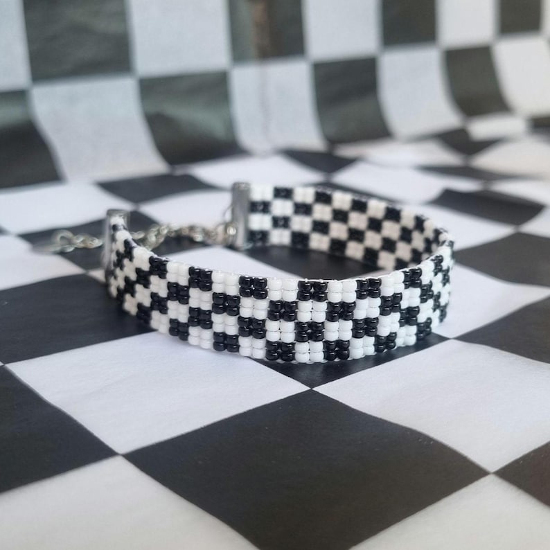 Puede incluir: Un brazalete de cuentas de color negro y blanco con un cierre plateado. El brazalete est&aacute; fotografiado sobre una superficie de cuadros negros y blancos.