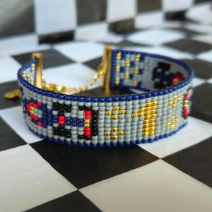 Puede incluir: Un brazalete de cuentas con un patrón geométrico en tonos de azul, gris, rojo, dorado y negro. El brazalete tiene un cierre de cadena dorada.