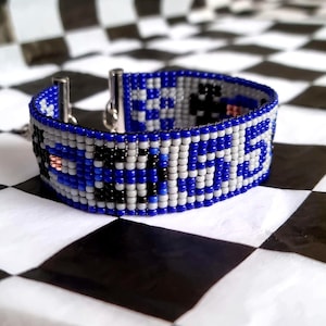 Puede incluir: Un brazalete de cuentas azules, blancas y negras con un cierre plateado. El brazalete presenta un patrón a cuadros y un diseño que se asemeja a un tablero de ajedrez.