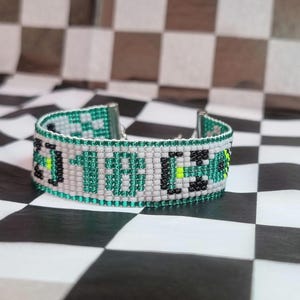 Op de afbeelding: Een kralenarmband met het woord "1988" in witte, groene, zwarte en gele kralen. De armband is geplaatst tegen een zwart-witte geblokte achtergrond. De armband heeft een zilveren sluiting.