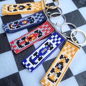 Puede incluir: Cinco llaveros rectangulares hechos de pequeñas cuentas de vidrio de colores en un patrón tejido. Cada llavero presenta un diseño geométrico único y una anilla de metal en la parte superior. Los colores incluyen amarillo, azul marino, rojo y blanco, con detalles en negro.